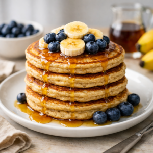 Fluffy banana oat pancakes stacked on a plate with fresh banana slices, blueberries, and maple syrup, healthy homemade breakfast recipe